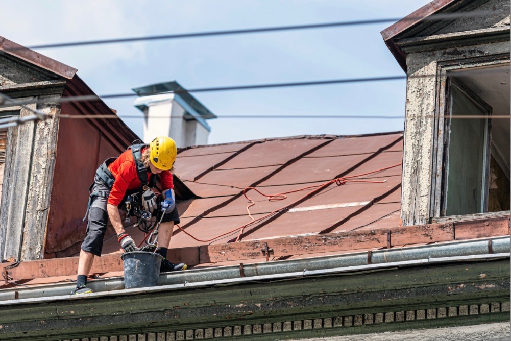 Pracownik w kasku i uprzęży zabezpieczającej, stojący na krawędzi dachu z blachy, z wiadrem w ręku, przygotowuje się do pracy na tle budynku z widocznym kominem i oknem z łuszczącą się farbą.