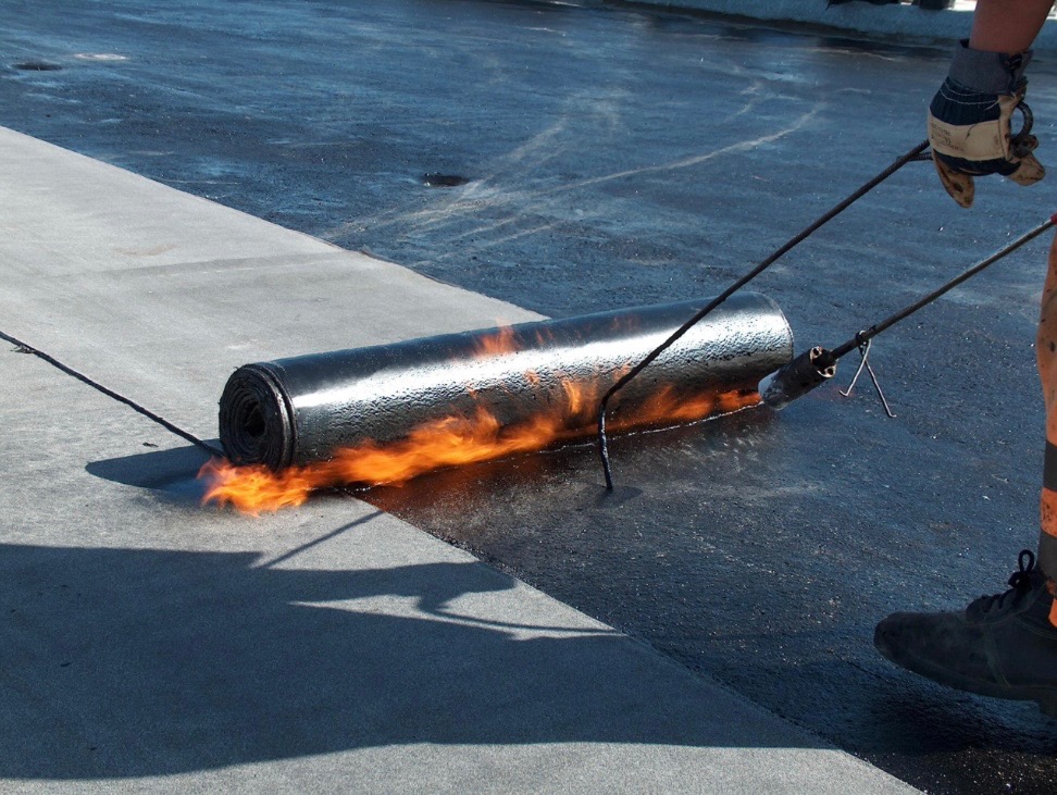 Pracownik z palnikiem w ręku rozgrzewa bitumiczną rolkę papy na płaskim dachu, widoczny płomień i świeżo pokryta powierzchnia.