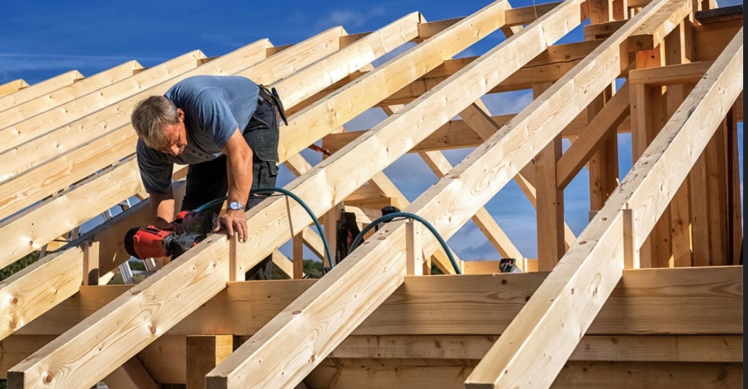 Dekarz mocuje drewniane elementy konstrukcji dachu za pomocą narzędzia pneumatycznego, widoczny fragment niebieskiego nieba w tle.