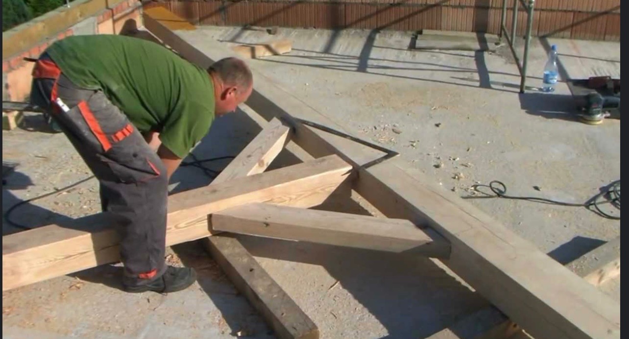 Monter w zielonej koszulce pochyla się nad drewnianą konstrukcją dachu w trakcie budowy, widoczne elementy więźby dachowej i narzędzia na betonowej powierzchni.