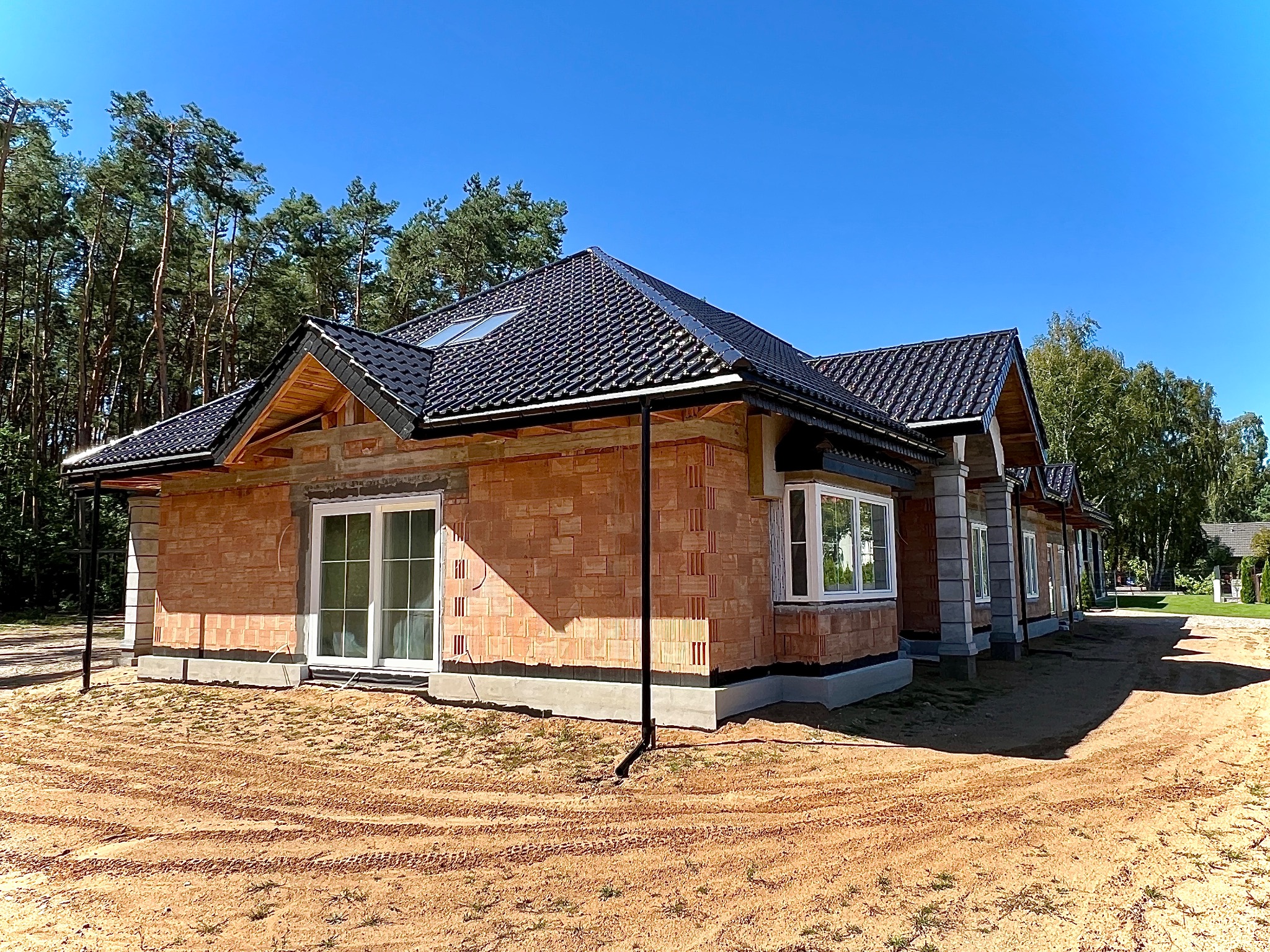 Dom jednorodzinny w trakcie budowy z czarnym dachem z dachówki ceramicznej, widoczne drewniane elementy konstrukcji dachu, okna PCV, ściany z cegły ceramicznej porotherm, teren wokół domu jeszcze...