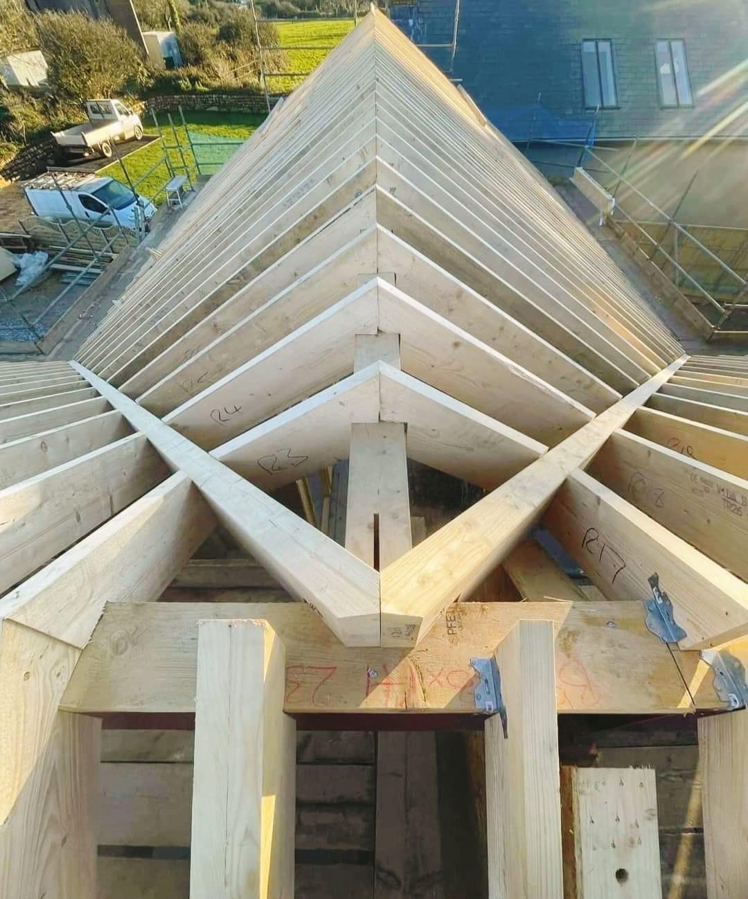 Konstrukcja drewniana dachu w trakcie budowy, widoczne krokwie i inne elementy nośne, oznaczone numerami, w tle budynek i zieleń.