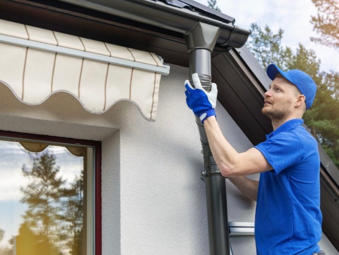 Mężczyzna w niebieskiej koszulce i czapce montuje szarą rynnę spustową na tle białej ściany budynku z markizą i widocznym fragmentem dachu.