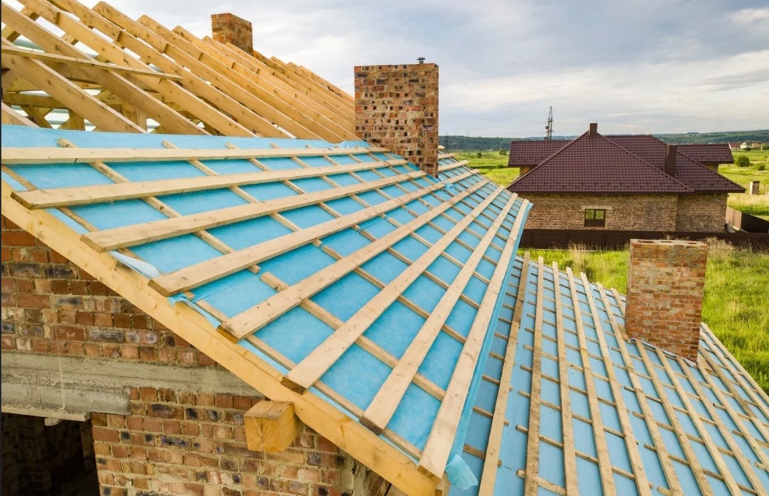 Konstrukcja dachu z drewnianymi krokwiami i niebieską membraną hydroizolacyjną, widoczny komin z cegły i dom w tle.