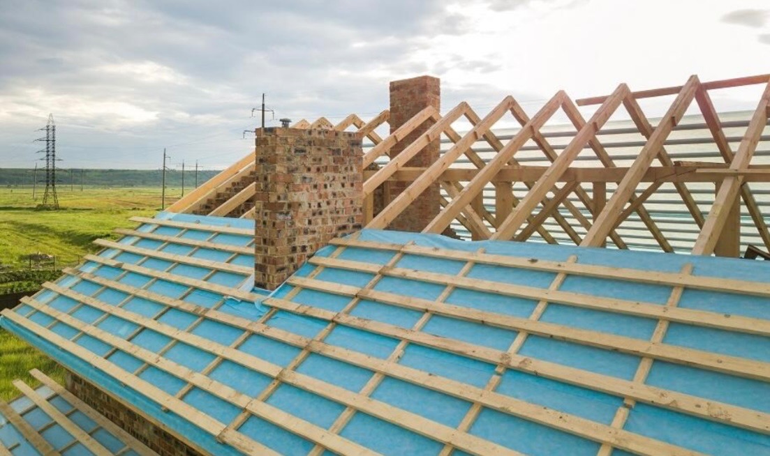 Konstrukcja drewniana dachu z widocznym kominem z cegły oraz niebieską membraną dachową, w tle słupy wysokiego napięcia i zielone pola.