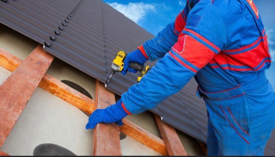 Pracownik w niebiesko-czerwonym kombinezonie mocuje ciemnoszare blachy faliste do drewnianej konstrukcji dachu za pomocą wkrętarki.