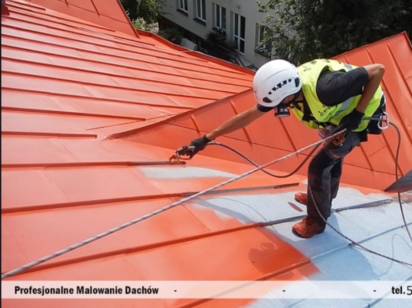 Pracownik w kasku i masce maluje natryskowo blaszany dach na pomarańczowo, zabezpieczony linami, widoczne częściowo pomalowane fragmenty na szaro.
