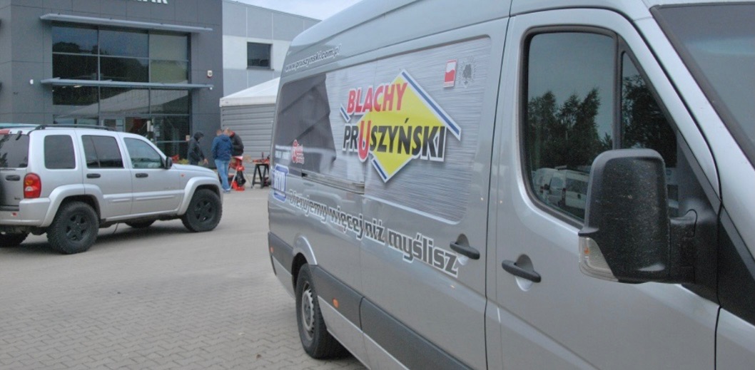 Srebrny bus z logo firmy dekarskiej Blachy Pruszyński na tle budynku firmy i zaparkowanego samochodu terenowego.