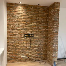 Ściana z cegły dekoracyjnej w salonie, z zamontowanym uchwytem na telewizor i gniazdkami elektrycznymi, widoczne kable i butelka wody na podłodze.