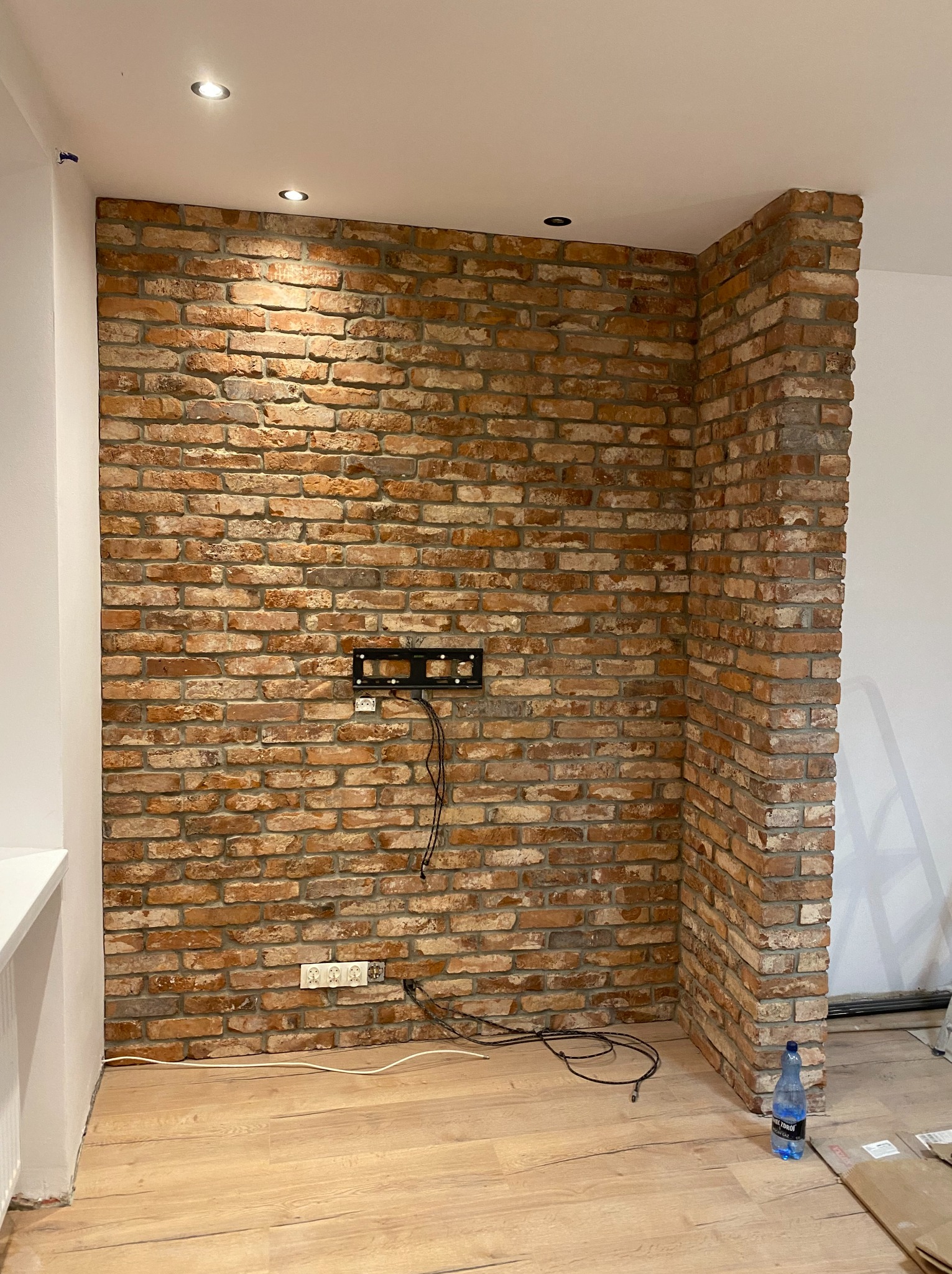 Ściana z cegły dekoracyjnej w salonie, z zamontowanym uchwytem na telewizor i gniazdkami elektrycznymi, widoczne kable i butelka wody na podłodze.