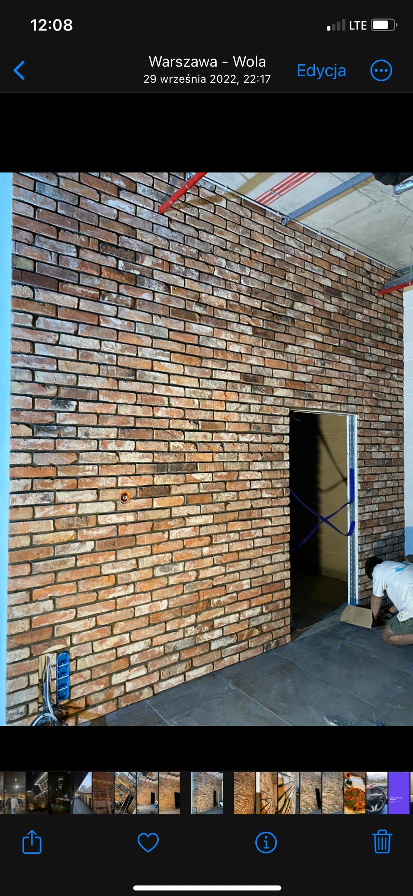 Ściana wykończona cegłą klinkierową z widoczną instalacją elektryczną i otworem drzwiowym w trakcie montażu ościeżnicy, pracownik w tle.