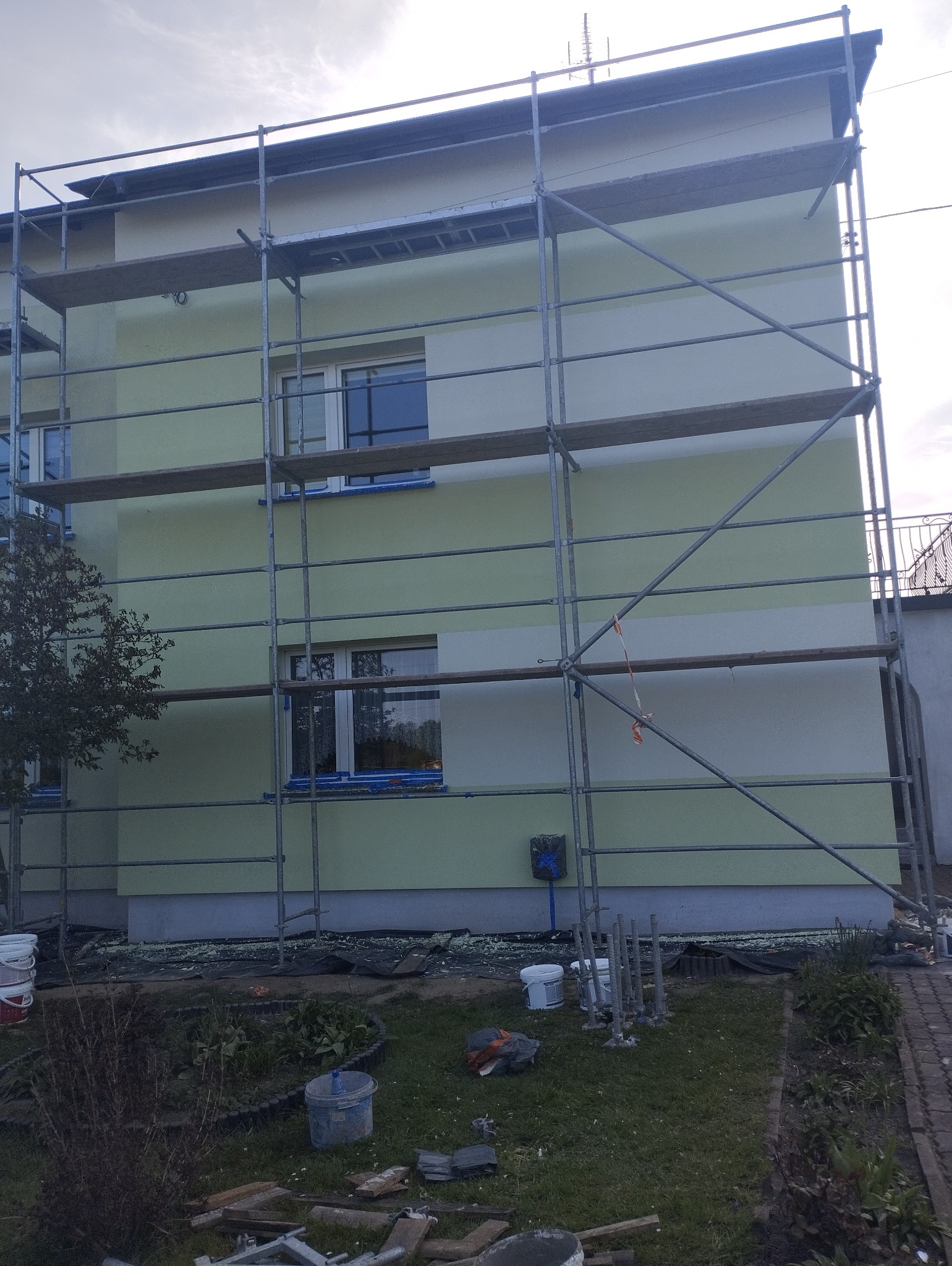 Dwukolorowa elewacja budynku w trakcie odświeżania, widoczne rusztowania, zabezpieczone okna taśmą malarską i resztki materiałów na trawniku.