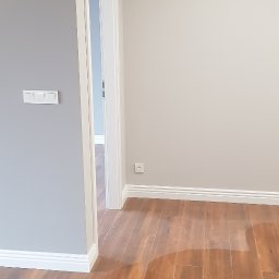 RAD-BUD Radosław Czubasiewicz - Pomalowane na szaro ściany, drewnopodobna podłoga i białe listwy przypodłogowe w pustym pokoju po remoncie, widok na otwarte drzwi do kolejnego pomieszczenia.