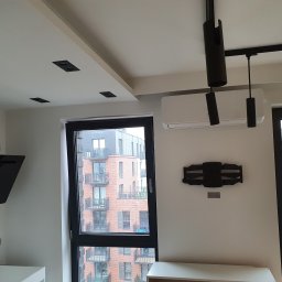 RAD-BUD Radosław Czubasiewicz - Nowoczesne wnętrze mieszkania z czarnymi reflektorami na szynie, klimatyzacją, dekoracją ścienną i widokiem na nowy budynek mieszkalny za oknem.