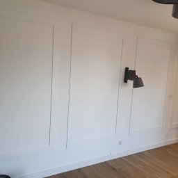 RAD-BUD Radosław Czubasiewicz - Biała ściana z dekoracyjnymi panelami w ramkach, drewniana podłoga, minimalistyczna lampa ścienna i oświetlenie sufitowe.