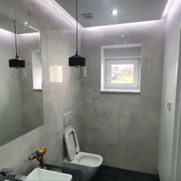 RAD-BUD Radosław Czubasiewicz - Szara łazienka z podświetleniem LED, wiszącą lampą z czarnym kloszem, umywalką i toaletą, w trakcie montażu widoczna wiertarka na umywalce.