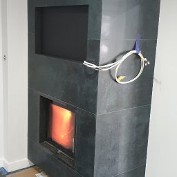 RAD-BUD Radosław Czubasiewicz - Nowoczesny kominek obłożony ciemnymi płytami z kamienia, z widocznym płomieniem i otwartą wnęką powyżej, przewody elektryczne na ścianie, zabezpieczona podłoga folią i kartonem.