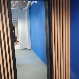 RAD-BUD Radosław Czubasiewicz - Drewniane lamele pionowe na ścianie z niebieskim akcentem, widok przez czarną ramę na wnętrze biura z szarym dywanem i biało-niebieską ścianą.