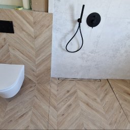 RAD-BUD Radosław Czubasiewicz - Nowoczesna łazienka z drewnopodobnymi płytkami ułożonymi w jodełkę na podłodze i ścianie, podwieszana toaleta z czarnym przyciskiem, szara ściana prysznicowa z czarną armaturą i odpływem liniowym.