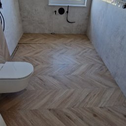 RAD-BUD Radosław Czubasiewicz - Wykończona łazienka z drewnopodobną podłogą ułożoną w jodełkę, białą wiszącą toaletą, jasnymi płytkami na ścianach i oknem z widokiem na zielone pole.