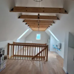 Poddasze po adaptacji z drewnianymi belkami stropowymi, oświetleniem industrialnym i drewnianą barierką, widoczne okno dachowe i instalacja elektryczna na ścianie.