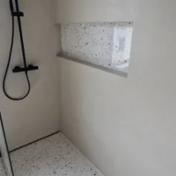 Nowoczesna łazienka z czarną armaturą prysznicową, tynkiem dekoracyjnym na ścianach i podłogą z lastryko oraz wnęką w ścianie wykończoną tym samym materiałem.