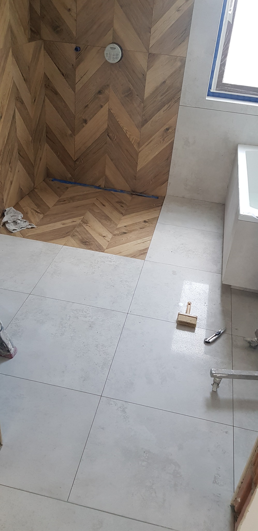 Układanie drewnopodobnych płytek w jodełkę na ścianie w Łambinowicach, w tle białe płytki podłogowe i fragment wanny, widoczny pędzel do fugowania i nóż do cięcia płytek.