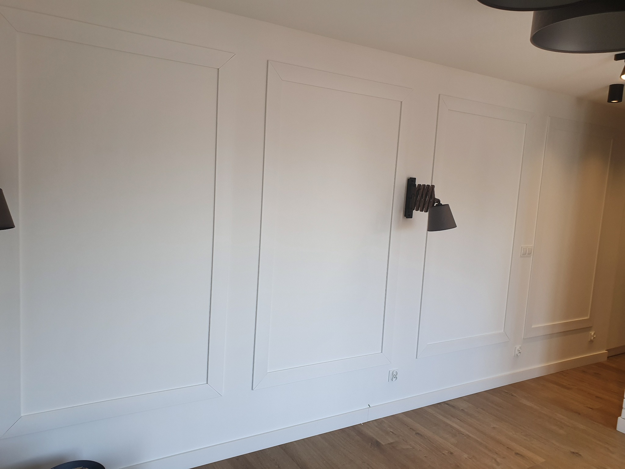 Biała ściana z dekoracyjnymi panelami w ramkach, drewniana podłoga, minimalistyczna lampa ścienna i oświetlenie sufitowe.