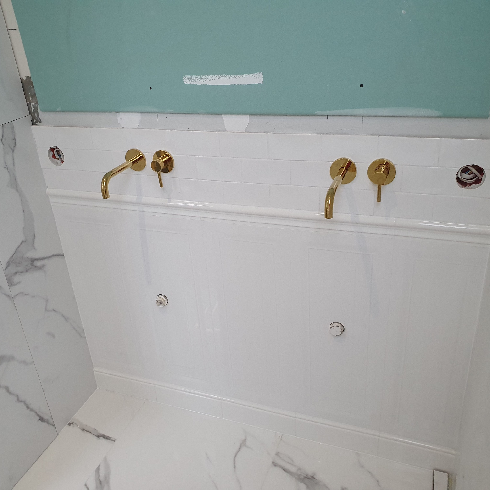 Biała, panelowa ściana w łazience z dwoma złotymi bateriami umywalkowymi, widoczne niedokończone otwory na instalacje elektryczne, marmurowe płytki podłogowe i fragment ściany z płyt...