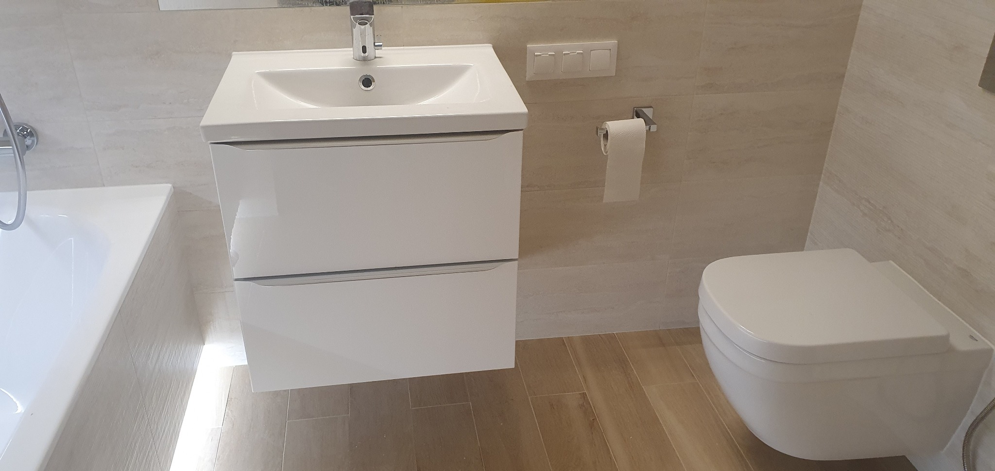 Nowoczesna, jasna łazienka z podwieszaną szafką umywalkową z dwoma szufladami, wiszącą toaletą i beżowymi płytkami imitującymi drewno na podłodze oraz ścianach.