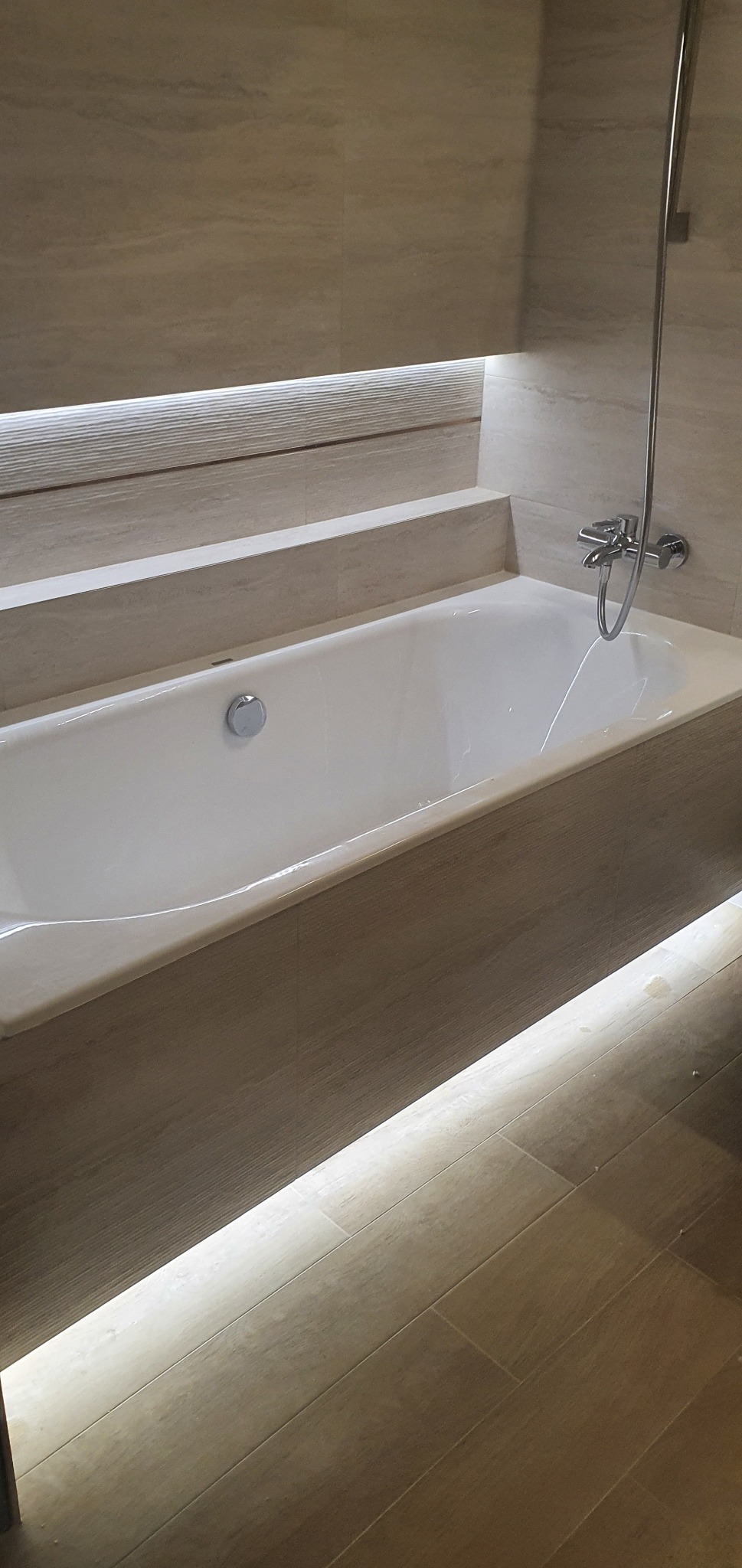 Nowoczesna wanna obudowana płytkami imitującymi drewno, z subtelnym podświetleniem LED od dołu i góry, chromowana bateria natryskowa zamontowana na ścianie z płytek.