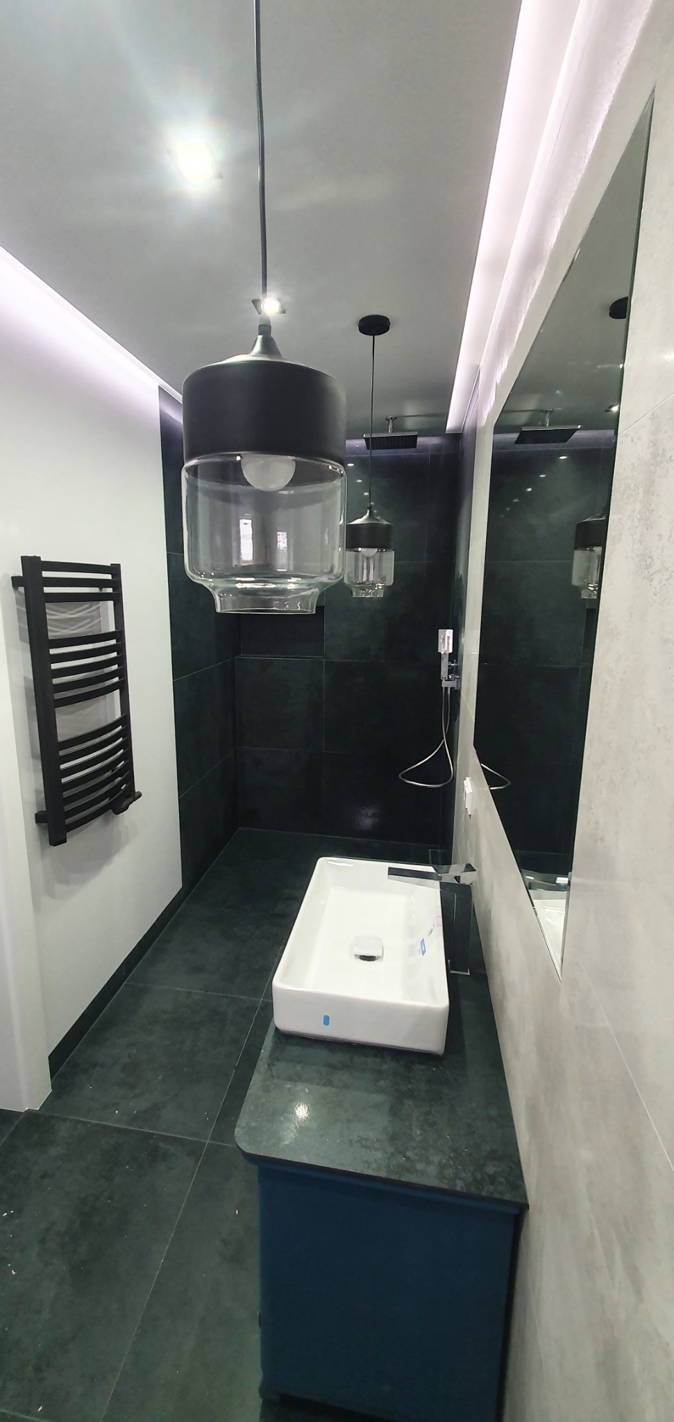 Nowoczesna łazienka z czarnymi płytkami, podwieszaną umywalką na szafce w kolorze petrol blue, dużym lustrem i designerskimi lampami wiszącymi z przezroczystymi kloszami.