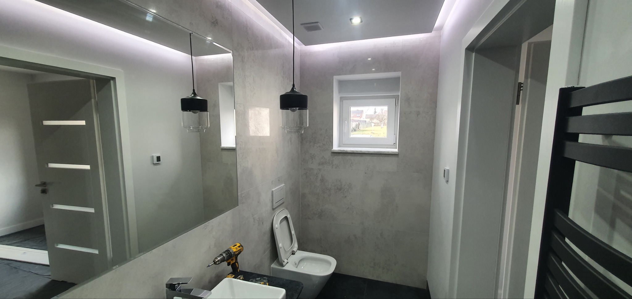Szara łazienka z podświetleniem LED, wiszącą lampą z czarnym kloszem, umywalką i toaletą, w trakcie montażu widoczna wiertarka na umywalce.