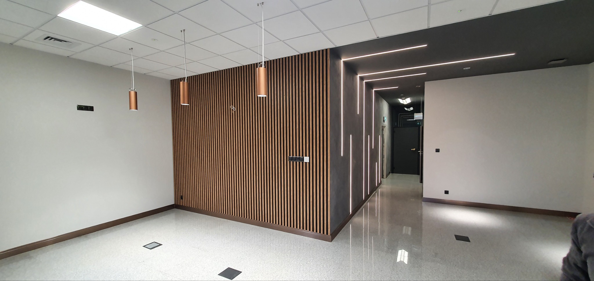 Nowoczesne wnętrze biurowe z drewnianą ścianą lamelową, oświetleniem LED i podwieszanymi lampami cylindrycznymi w kolorze miedzi.