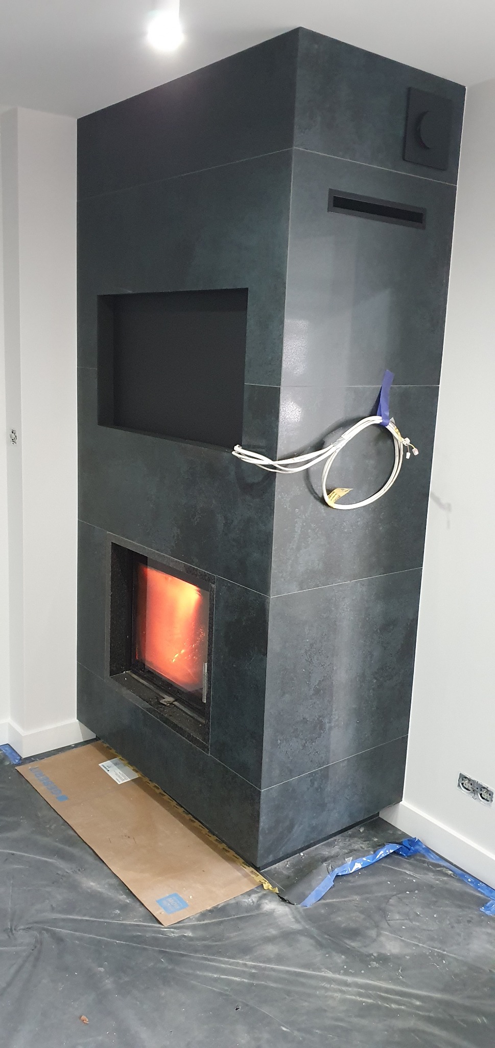 Nowoczesny kominek obłożony ciemnymi płytami z kamienia, z widocznym płomieniem i otwartą wnęką powyżej, przewody elektryczne na ścianie, zabezpieczona podłoga folią i kartonem.