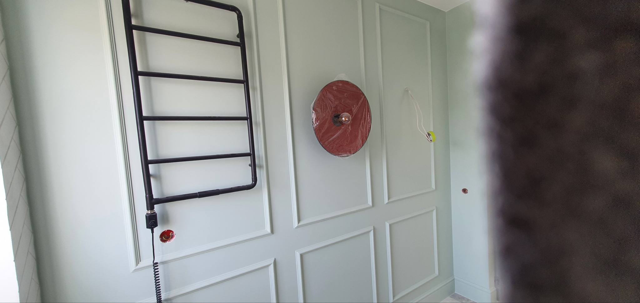 Ściana łazienki w trakcie prac wykończeniowych: grzejnik drabinkowy podłączony do instalacji elektrycznej, lampa ścienna w folii ochronnej, widoczne przewody elektryczne.