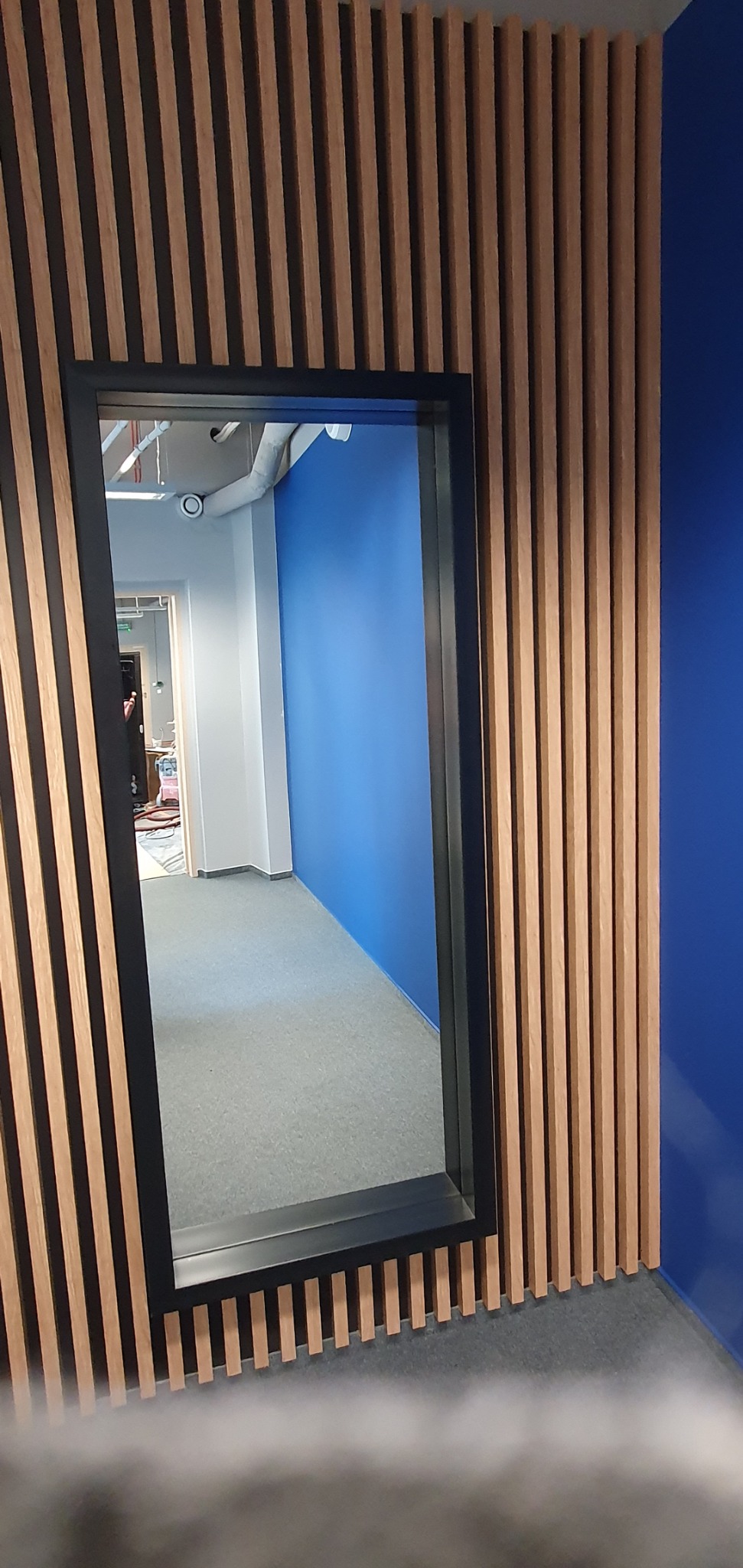 Drewniane lamele pionowe na ścianie z niebieskim akcentem, widok przez czarną ramę na wnętrze biura z szarym dywanem i biało-niebieską ścianą.