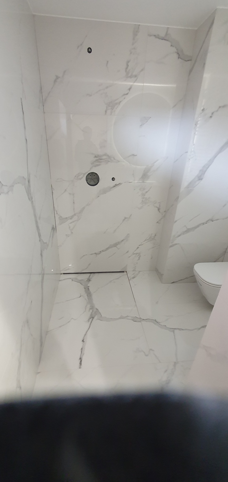 Wykończona łazienka z białymi płytkami imitującymi marmur, widoczny odpływ liniowy i podwieszana toaleta, brak kabiny prysznicowej.