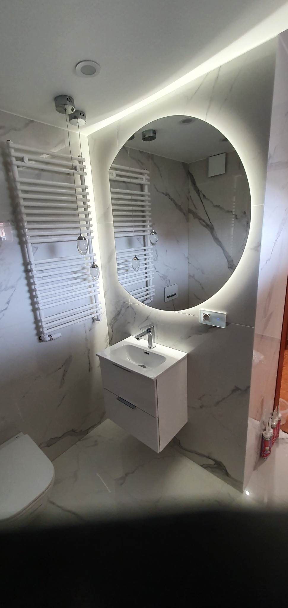 Nowoczesna łazienka z marmurowymi płytkami, okrągłym lustrem z podświetleniem LED, białą szafką pod umywalką z dwoma szufladami i grzejnikiem drabinkowym z dekoracyjnymi zawieszkami.
