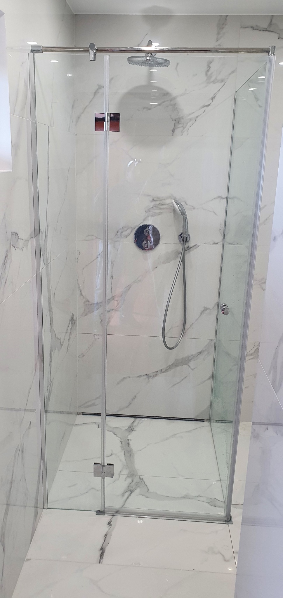Kabina prysznicowa z przezroczystego szkła z chromowanymi okuciami, zamontowana w łazience wyłożonej białymi płytkami imitującymi marmur, widoczna deszczownica i bateria.