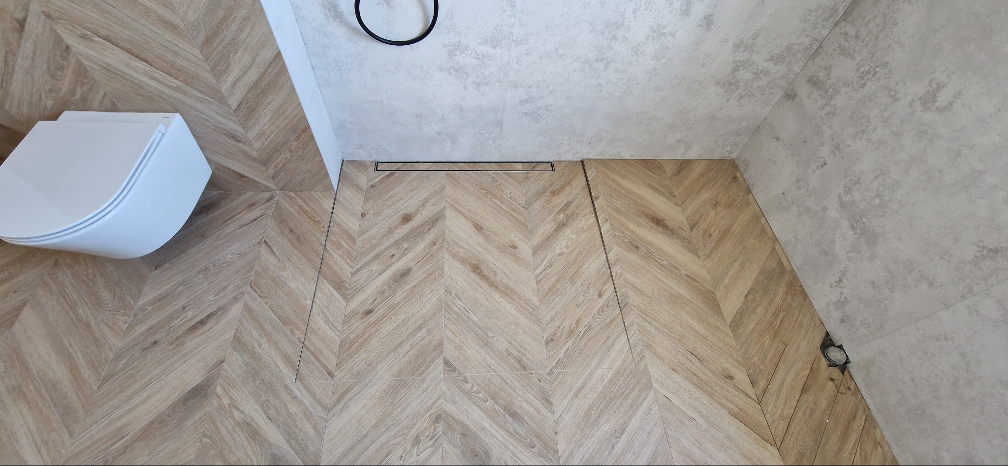 Wykończona łazienka z podłogą w jodełkę z drewnopodobnych płytek, odpływ liniowy i podwieszana toaleta z deską wolnoopadającą.