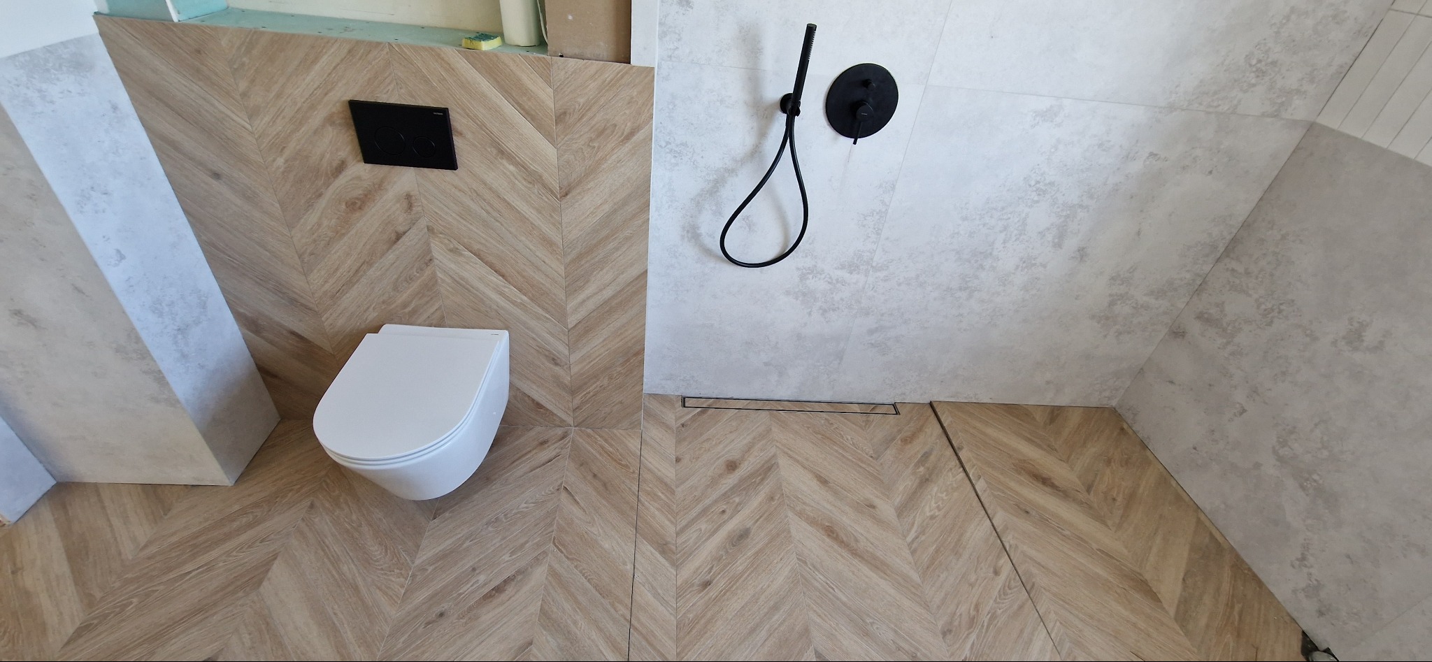 Nowoczesna łazienka z drewnopodobnymi płytkami ułożonymi w jodełkę na podłodze i ścianie, podwieszana toaleta z czarnym przyciskiem, szara ściana prysznicowa z czarną armaturą i odpływem liniowym.