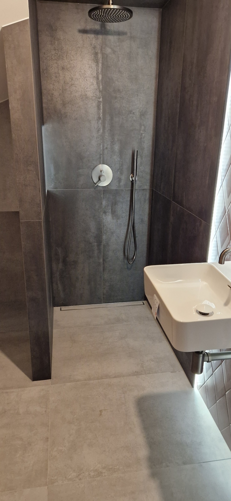 Nowoczesna łazienka z szarymi płytkami imitującymi beton, deszczownicą i minimalistyczną umywalką nablatową. Widoczny odpływ liniowy w podłodze.