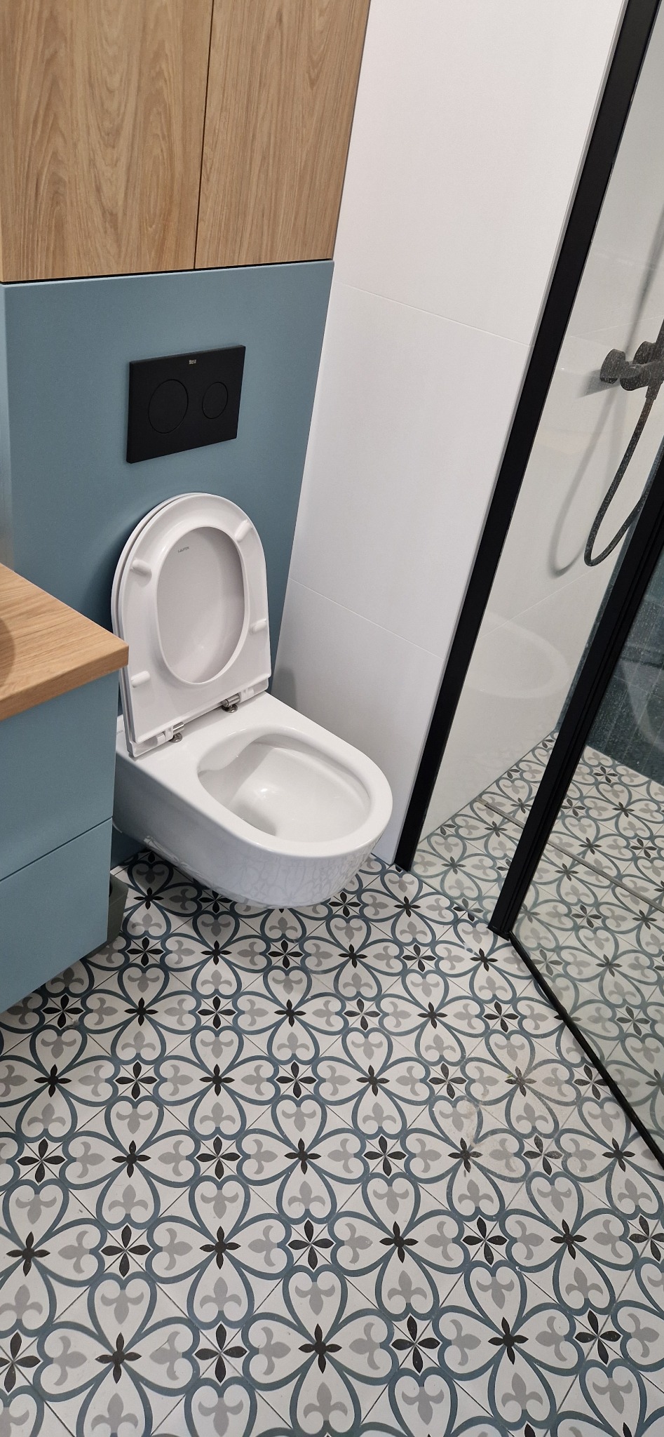 Nowoczesna toaleta wisząca marki Laufen z czarnym przyciskiem spłukującym na niebieskiej ścianie, wzorzyste płytki podłogowe i fragment kabiny prysznicowej z czarną ramą.