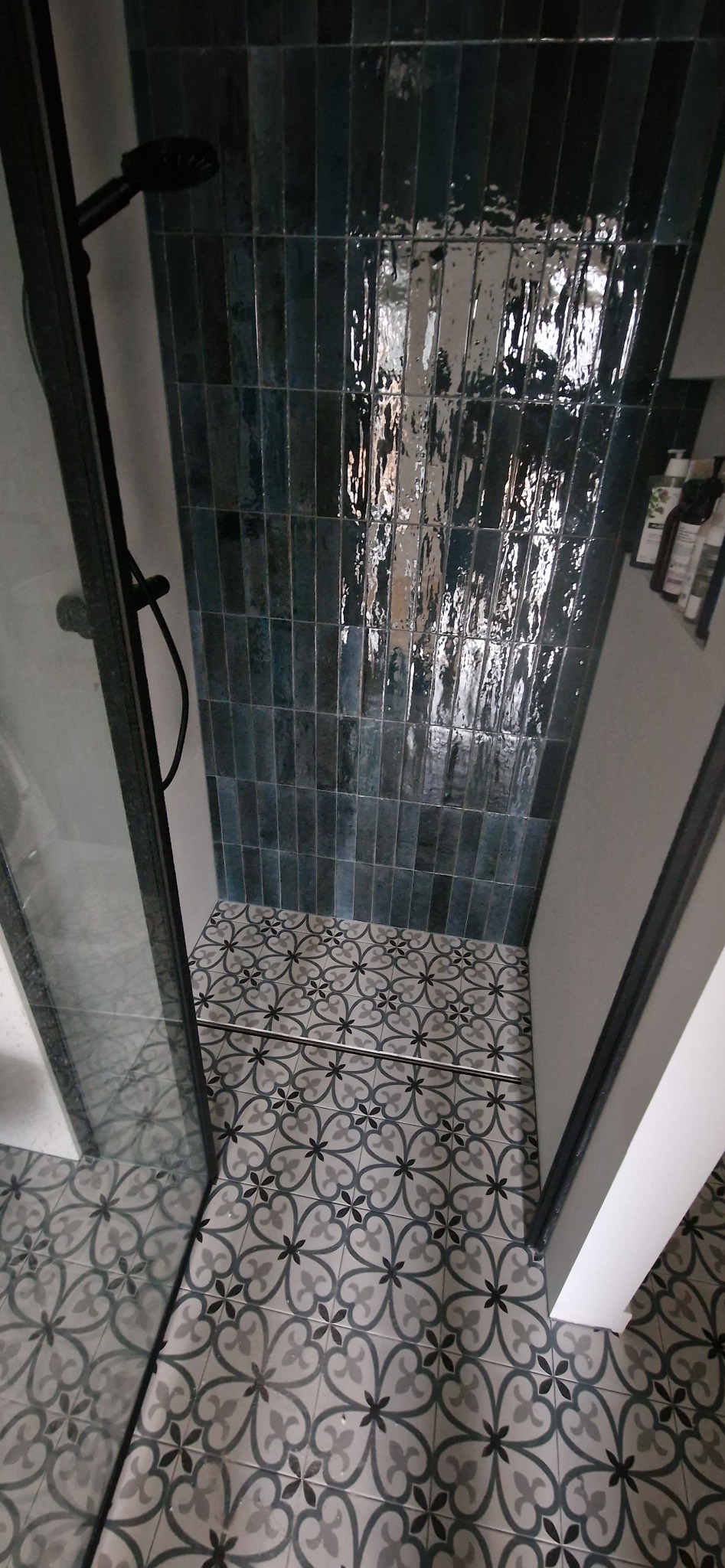 Nowoczesna kabina prysznicowa z ciemnymi, błyszczącymi płytkami ułożonymi pionowo na ścianie i wzorzystą, szaro-czarną podłogą, widok z góry.