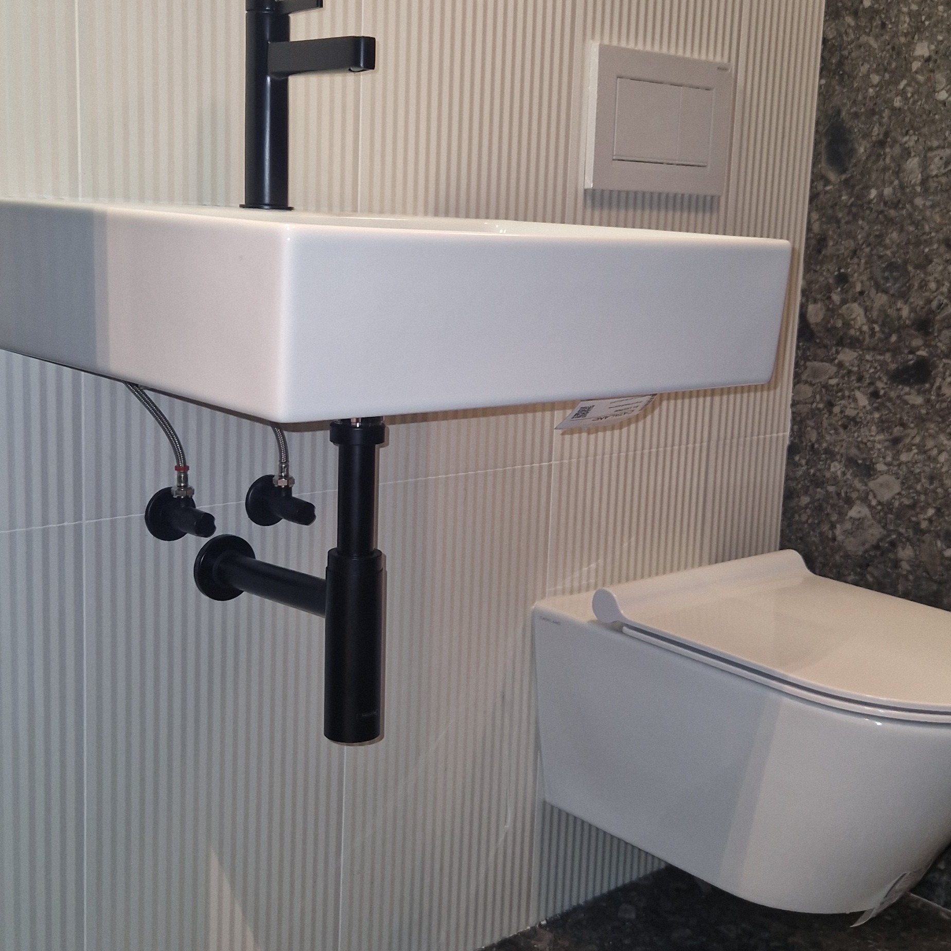 Biała, prostokątna umywalka z czarną baterią i odpływem na tle ściany z pionowymi paskami i wiszącej toalety.