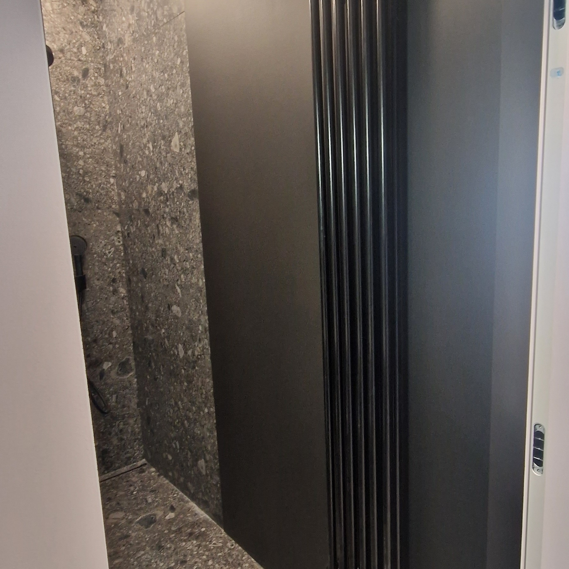 Fragment łazienki z szarymi płytkami imitującymi kamień, czarnym grzejnikiem drabinkowym i częściowo widoczną kabiną prysznicową z czarnymi elementami.