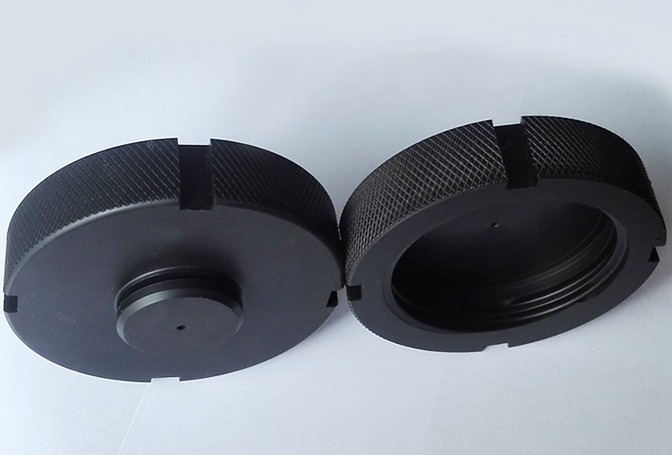 Dwie czarne, okrągłe, metalowe nakrętki z gwintem wewnętrznym i zewnętrznym, leżące obok siebie na białym tle, z widocznymi detalami obróbki skrawaniem.