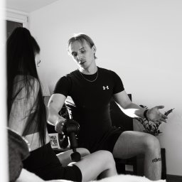 Jakub Kasiński - Mężczyzna w czarnym stroju sportowym z tatuażem na nodze demonstruje kobiecie użycie masażera wibracyjnego na udzie podczas sesji w pomieszczeniu.