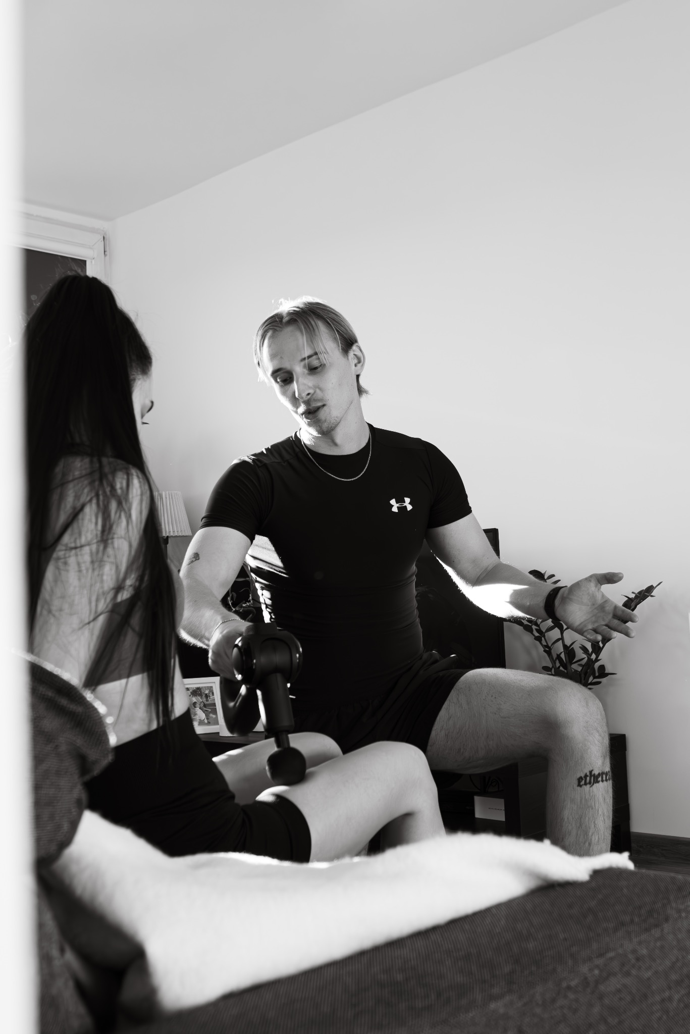 Mężczyzna w czarnym stroju sportowym z tatuażem na nodze demonstruje kobiecie użycie masażera wibracyjnego na udzie podczas sesji w pomieszczeniu.